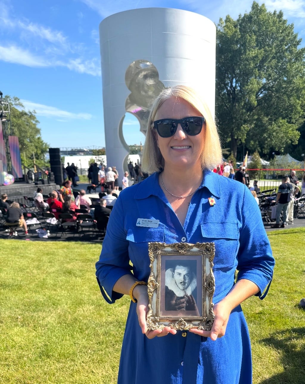 A woman in a blue dress holds a black and white photo while standing in front of the new 2SLGBTQ+ monument in Ottawa.