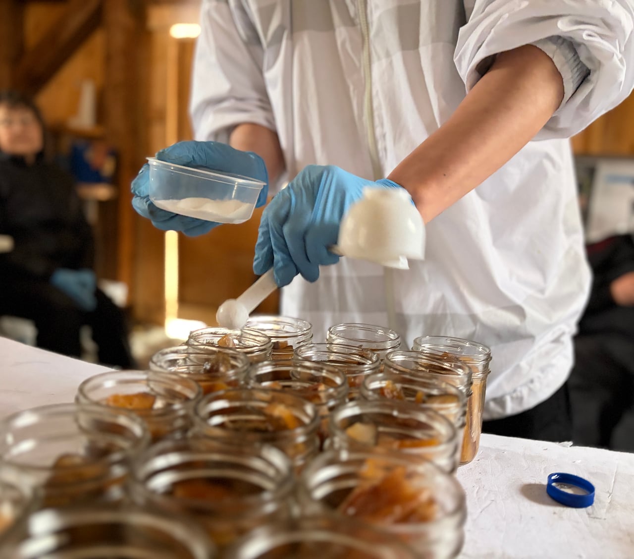 Jordan Cote-Germain, wearing a white jacket and blue gloves, adds salt to glass jars filled with smoked fish during a canning workshop.