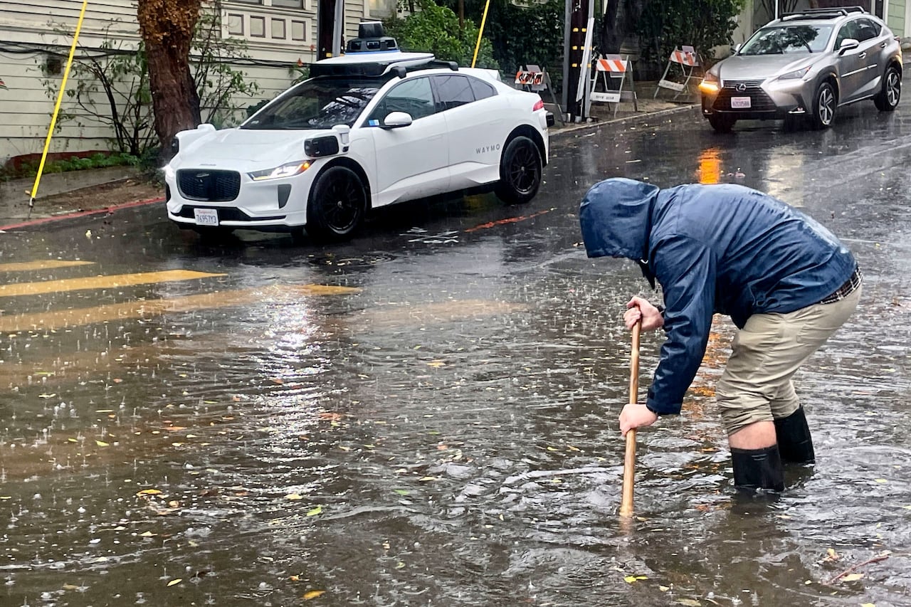 a man tries to clear a grate on a flooded street, as a waymo sits in the background