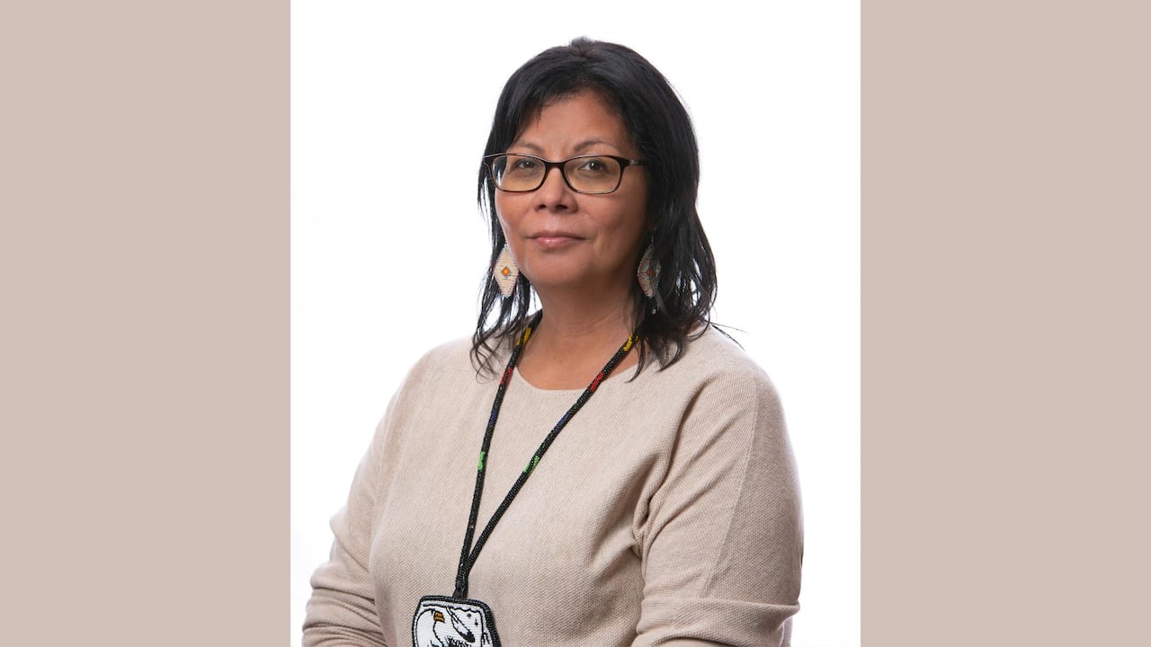 A headshot of a person wearing a beige shirt and a beaded medallion around their neck.