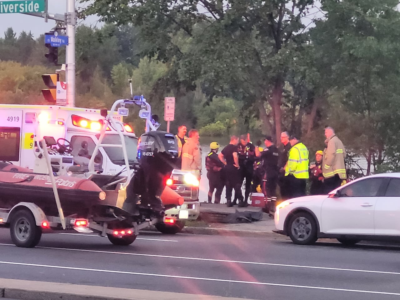 See emergency vehicles parked in a street and police officers and other first responders standing by a river.