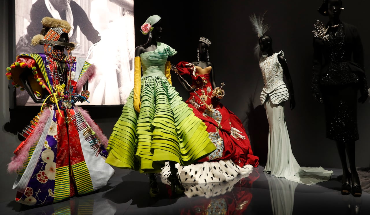 A few haute couture dresses are displayed on mannequins on a large black stage with a screen behind them.