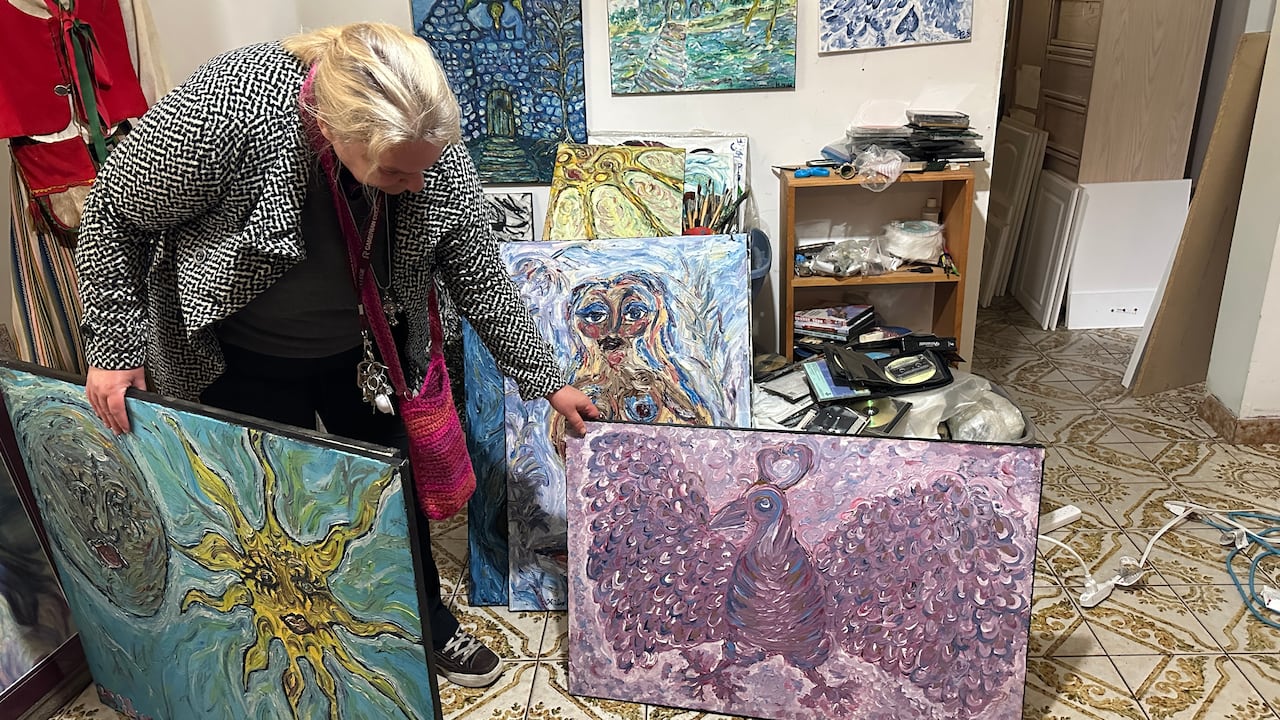 A woman with blonde hair holds up several canvasses with swirling figures.