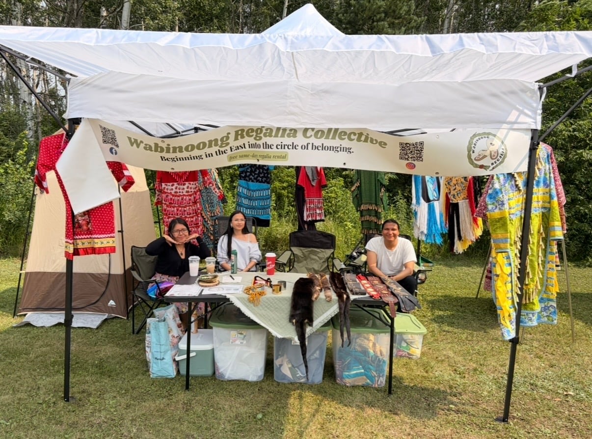 Three people sit in a vendor tent with "Wabinoong Regalia Collective" sign and regalia waiting for those who need it