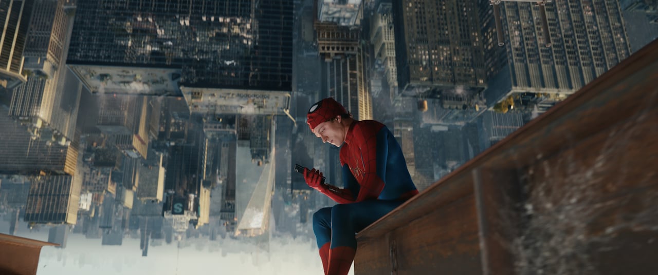 man in spiderman outfit sits atop a skyscraper looking at his phone , the backround is upside down.