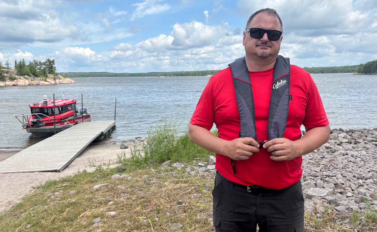 A man in a red shirt and sunglasses stands near the shore of a lake. A boat is moored to a dock in the background.