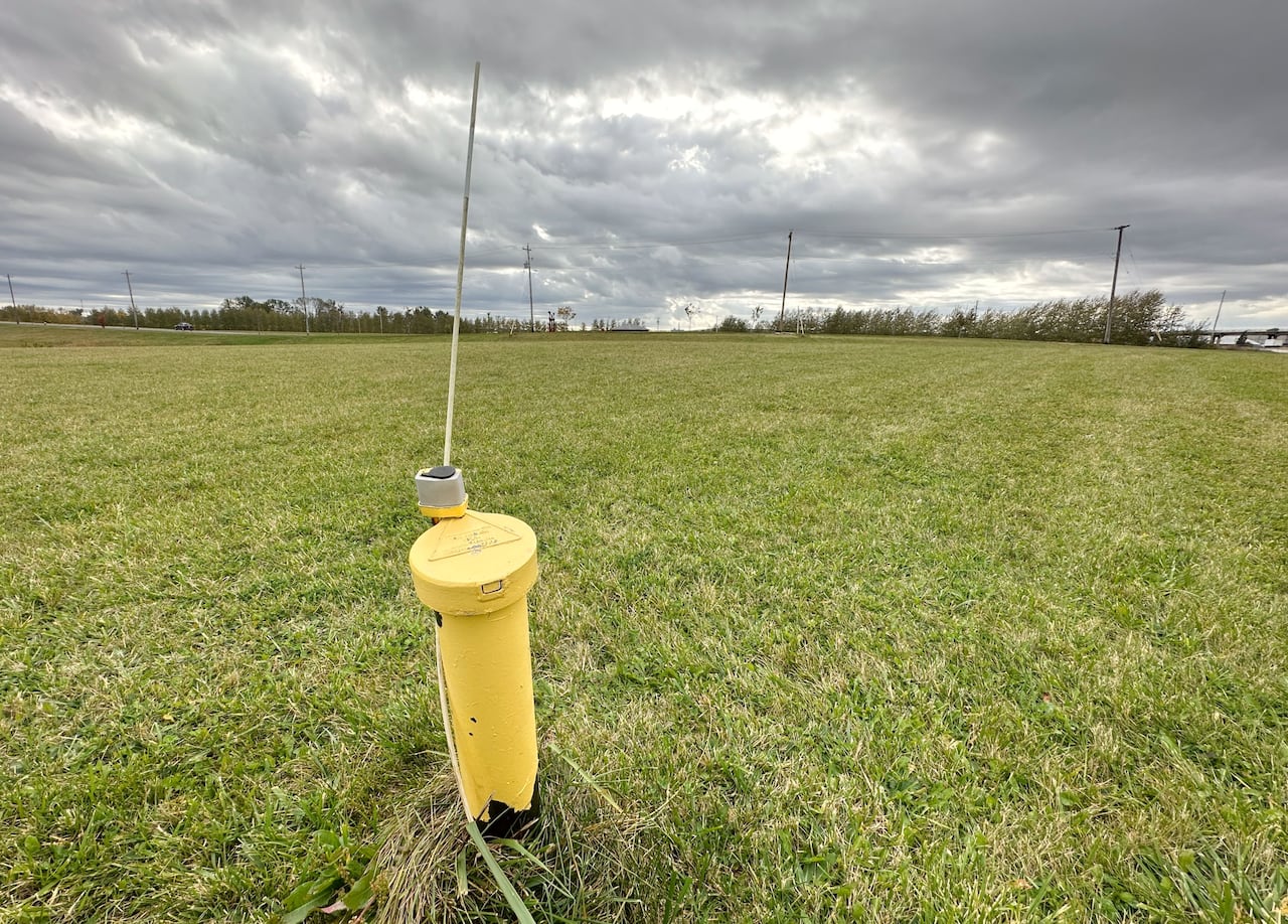 A yellow metal pipe with a metal cap on it and a metal pole sticking out the top is seen in the middle of a field of green grass.