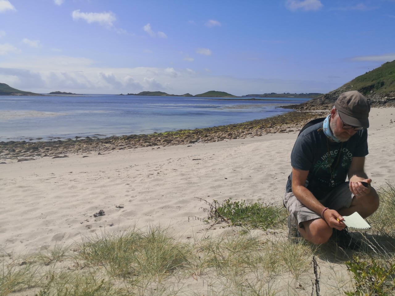 Man crouched on a beach writing in a notebook