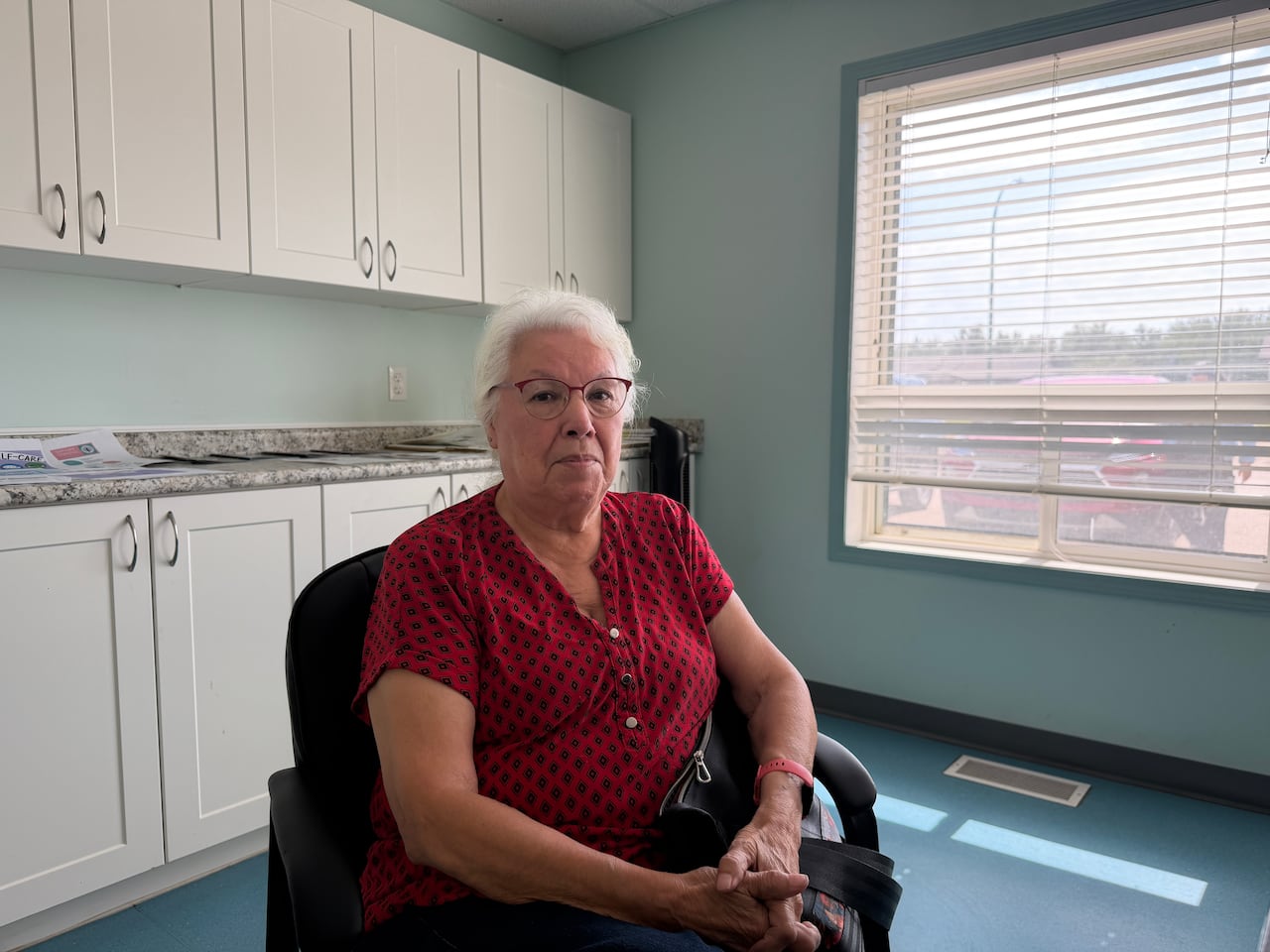 Elderly woman sits in a chair with a big window behind her.