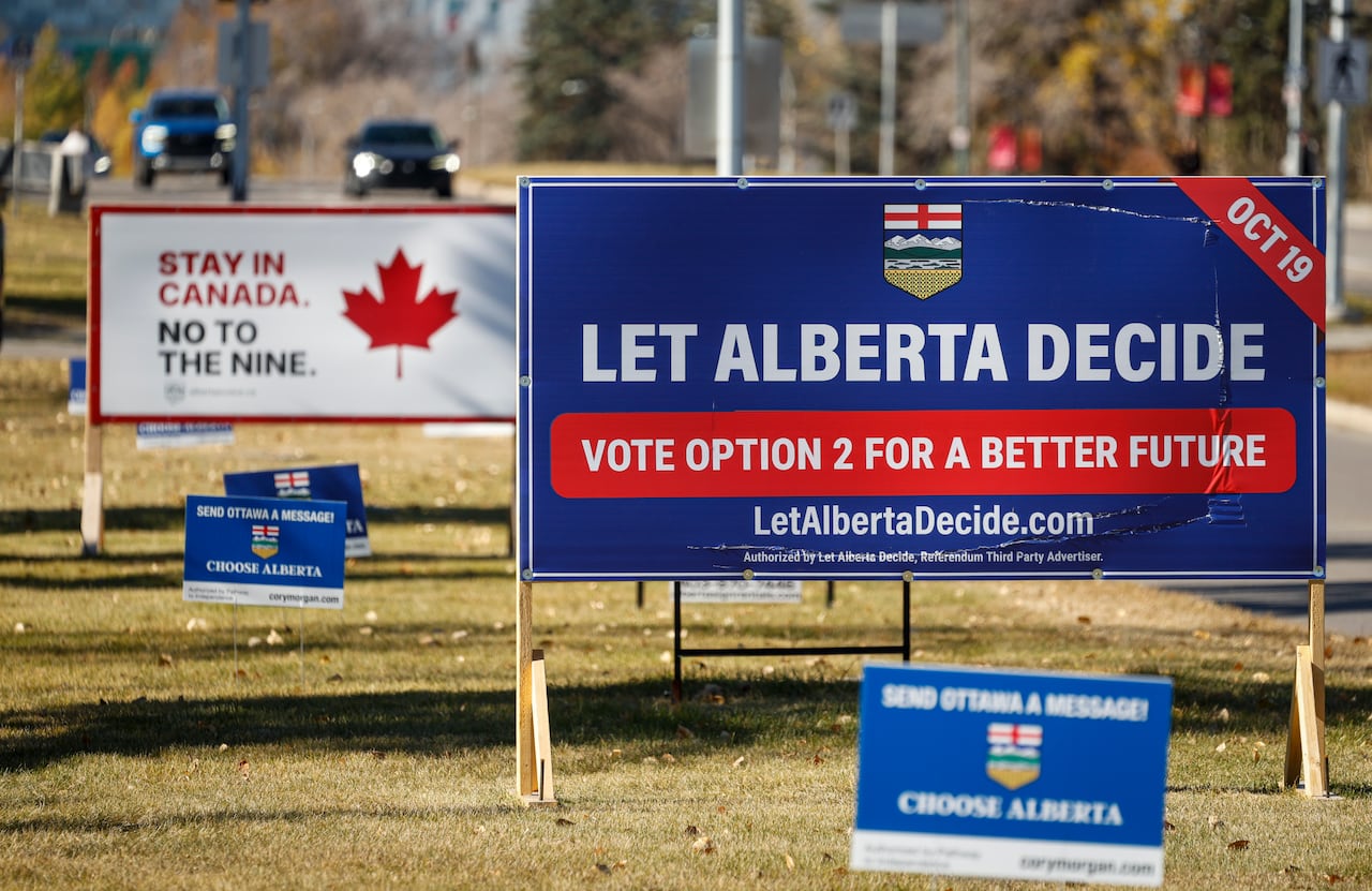 In front, a blue campaign sign that says Let Alberta Decide. Behind it, a white sign that says Stay in Canada. No to the Nine.