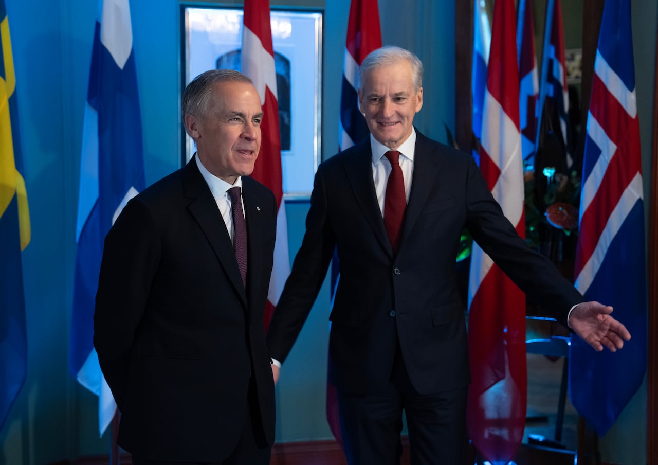 Two men in suits stand in front of the flags of several countries.