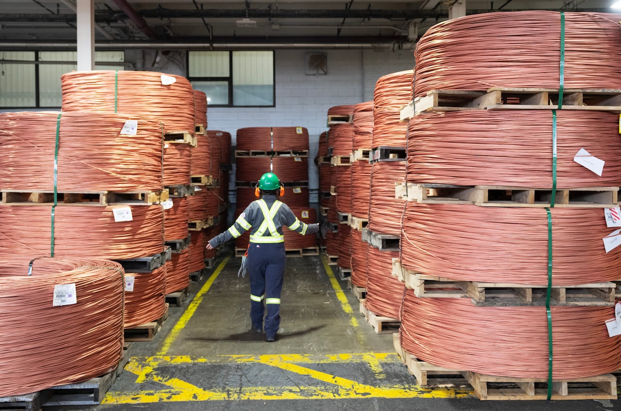 a person in a hard hat and safety gear walks between massive spools of copper wire in a warehouse
