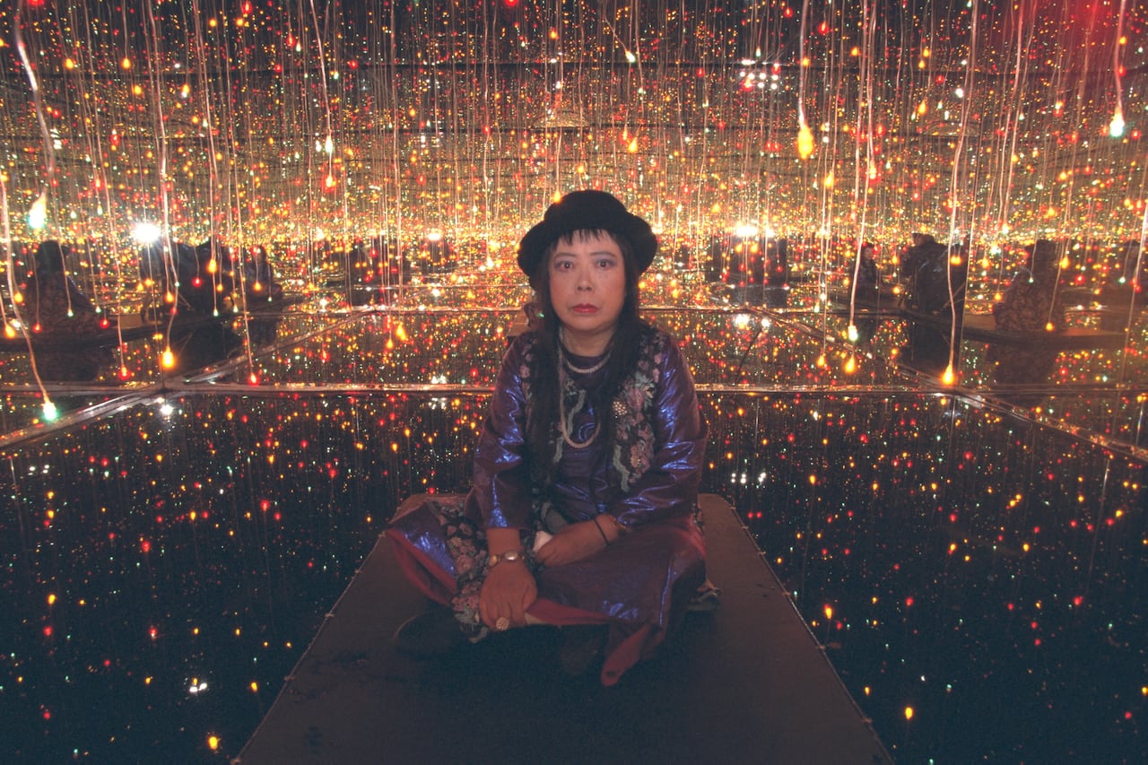A younger Yayoi Kusama sits cross legged in her installation surrounded by dazzling sparkly balls