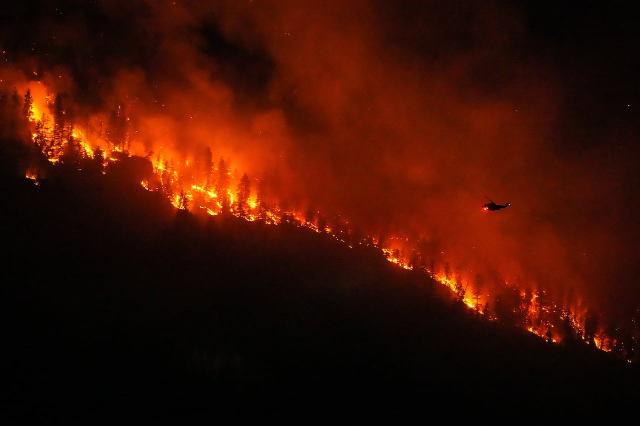 A helicopter flies above a hill fully engulfed in flames.