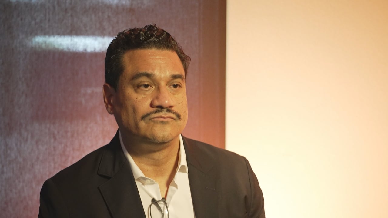 A man with a moustache wearing a black suit stands in front of a wall.