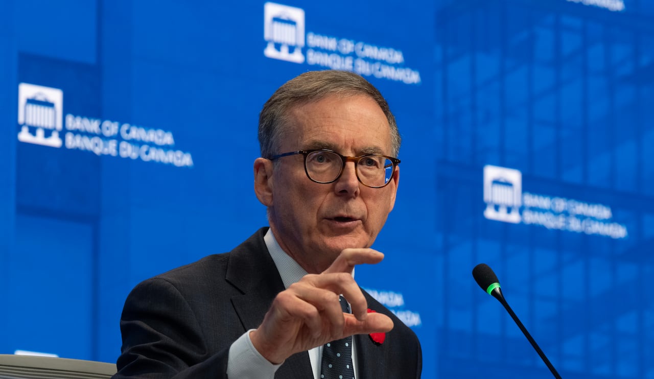 a man in a suit sits in front of a blue background, talking with his hands while answering a reporter's question
