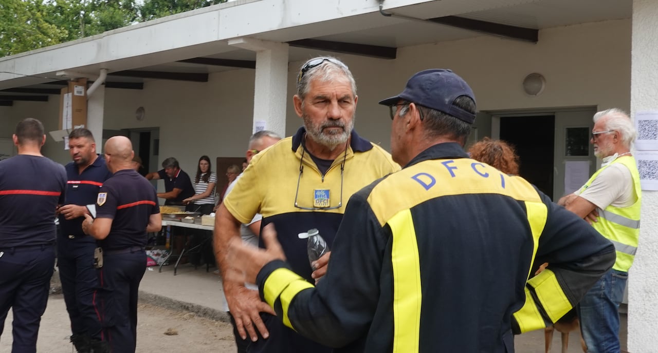 A man with grey hair and a yellow polo holds his hands on his hips and speaks to another man.