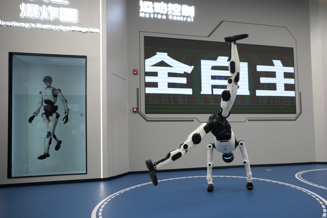 A white and black robot on its hands, one leg straight up, the other hanging low.