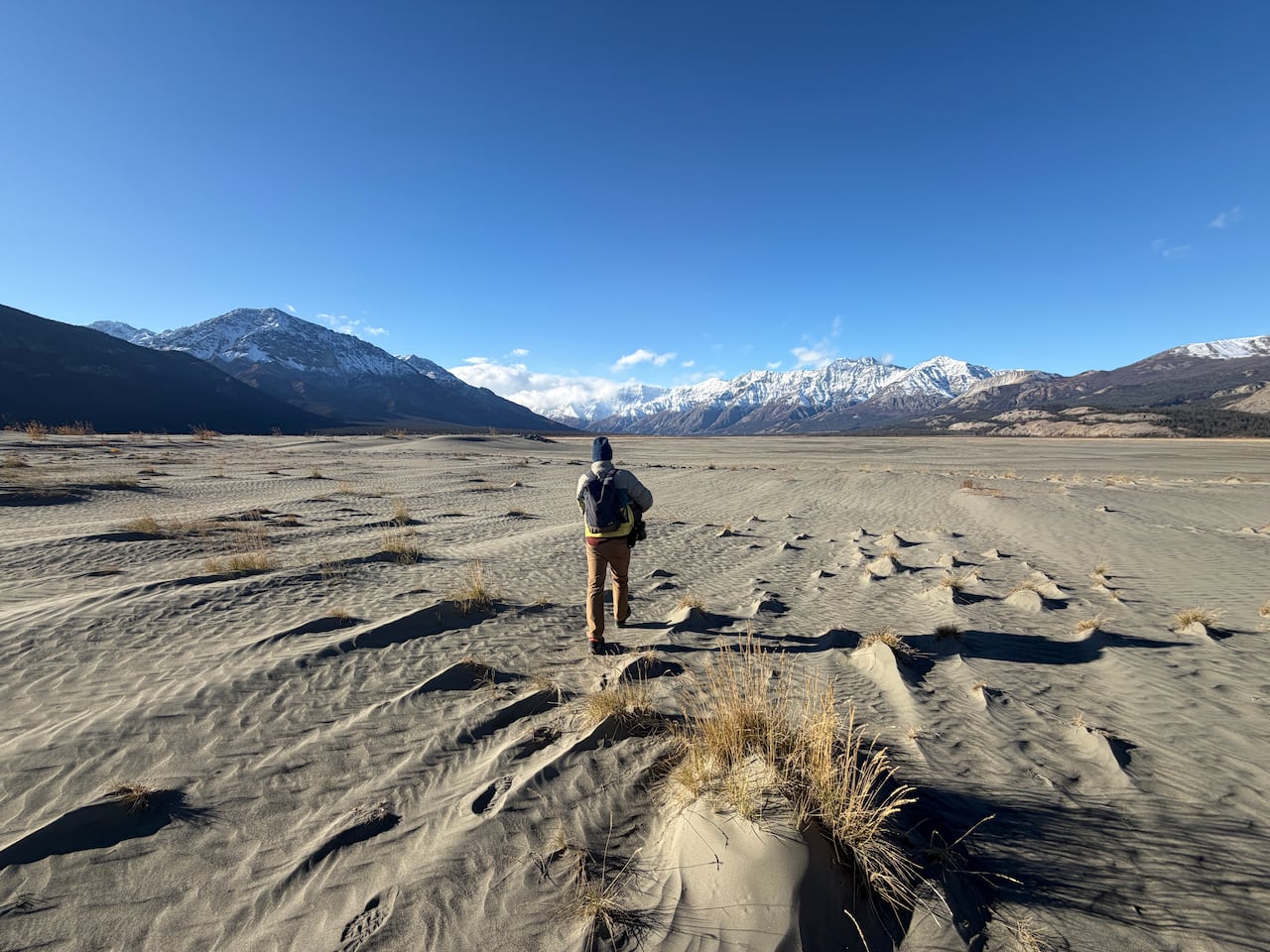 James King walks away from the camera across rippled grey sediment in the Ä'äy Chù Valley, with snow-covered mountains in the distance.