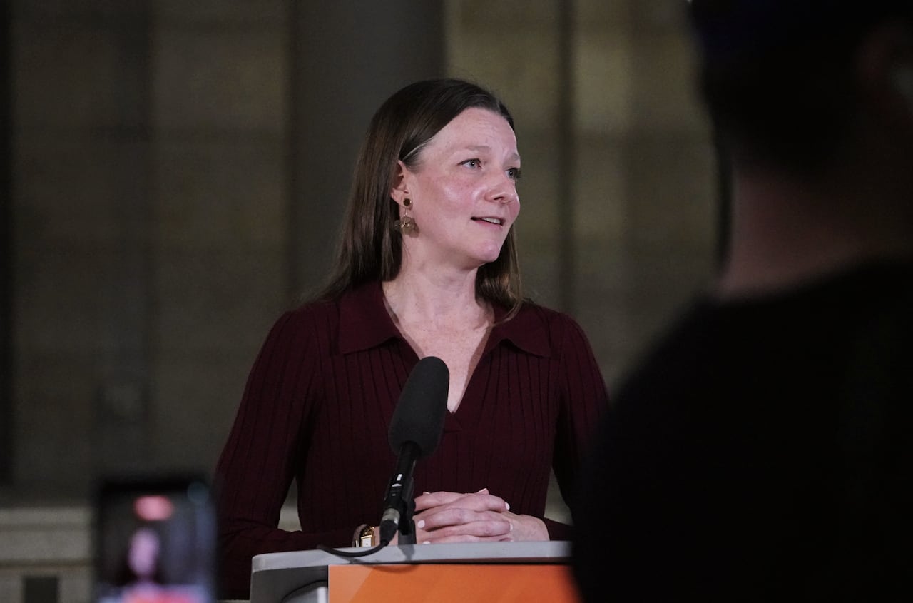 A woman with long brown hair in a maroon shirt clasps her hands on a podium while speaking into a microphone.