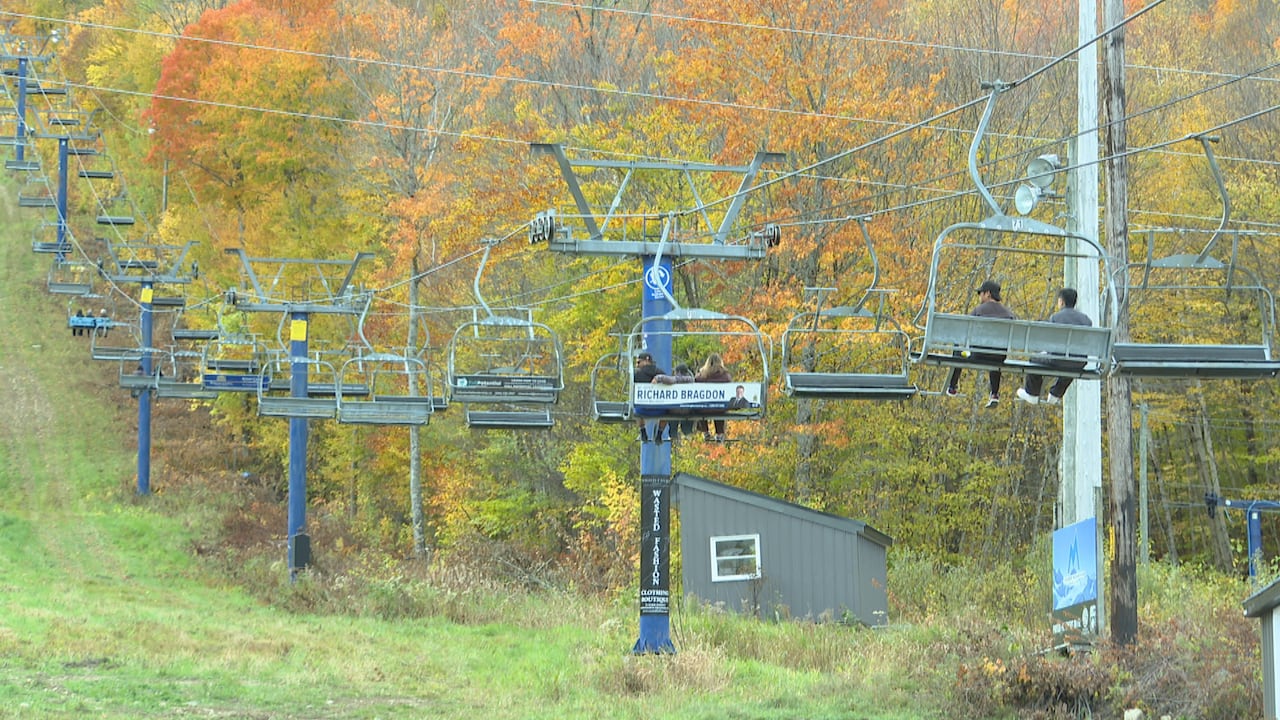 An operating ski chairlift carrying passengers up a grassy hill surrounded by dense forest with vibrant yellow and orange autumn foliage.