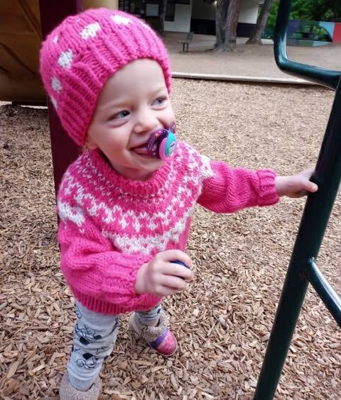 A toddler sucking a soother plays in a playground with wood chips on the ground. She is wearing a pink toque and pink sweater and is smiling.