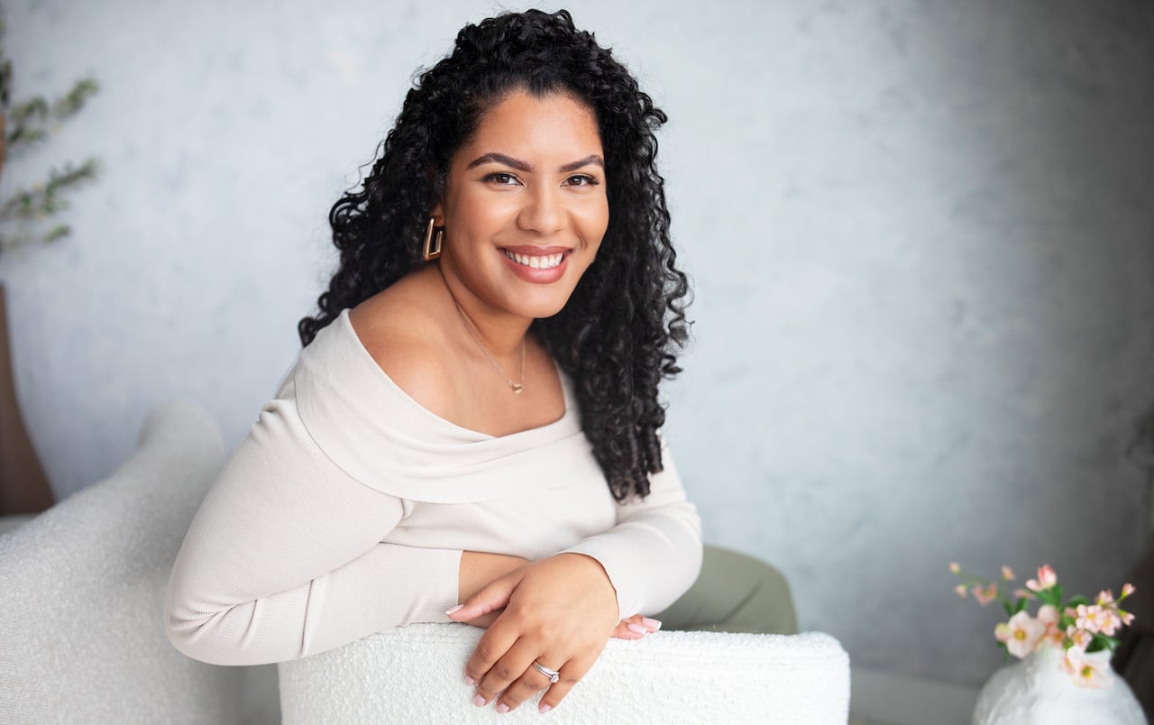 A Black woman in a white top smiles at the camera while leaning on the arm of an armchair.