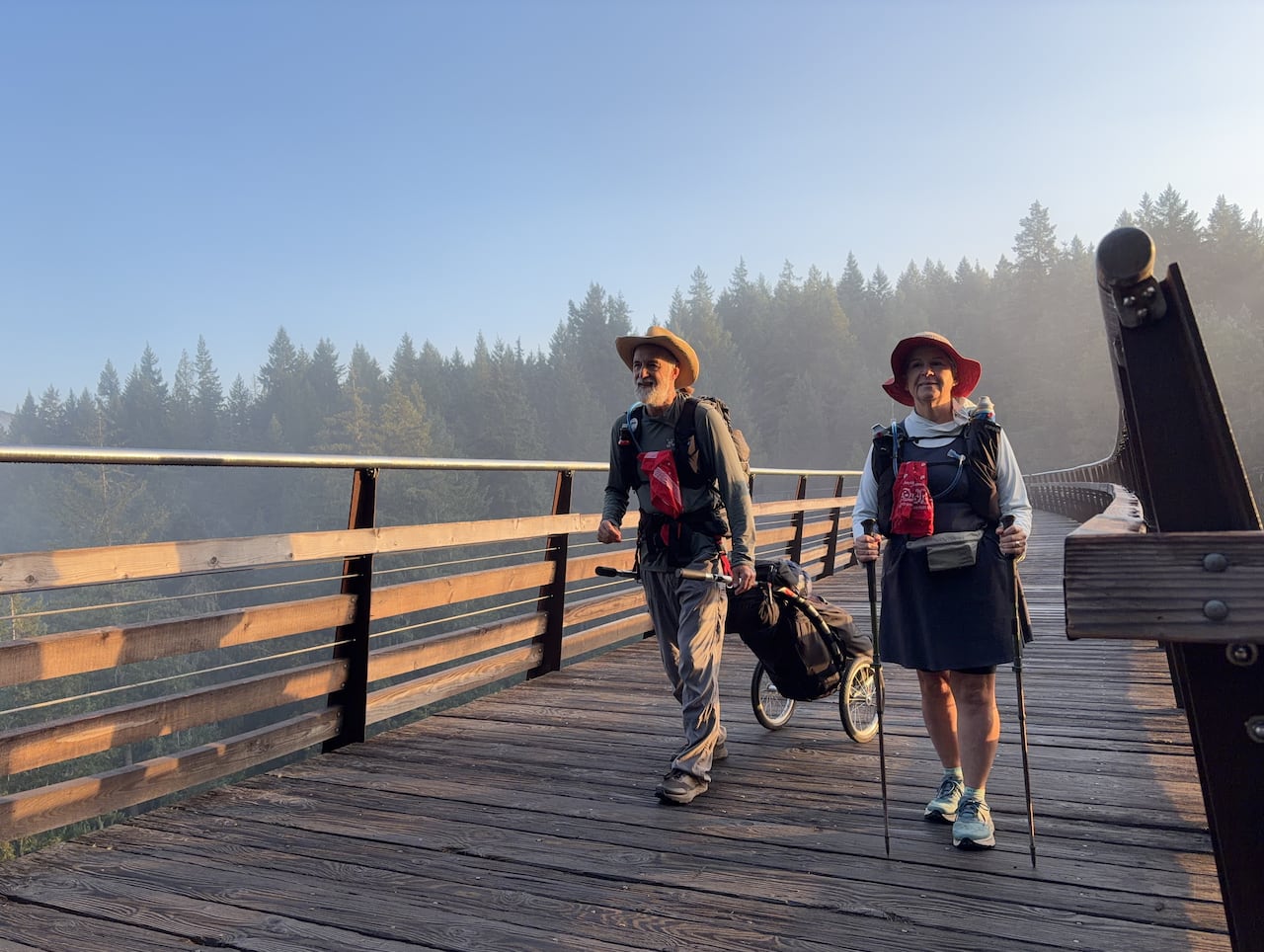 Two hikers wearing backpacks and sun hats walk across a trestle bridge with trekking poles.