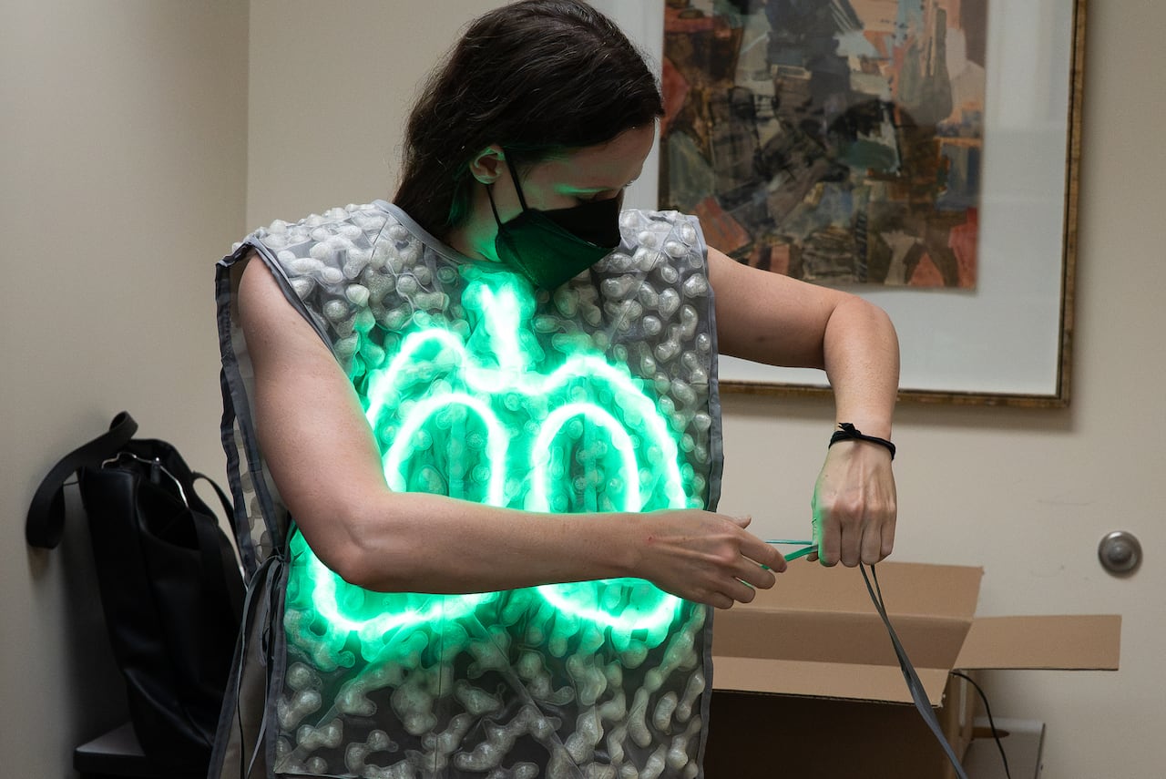 Woman with mask hooks up wires to a vest with illuminated green lungs on the front