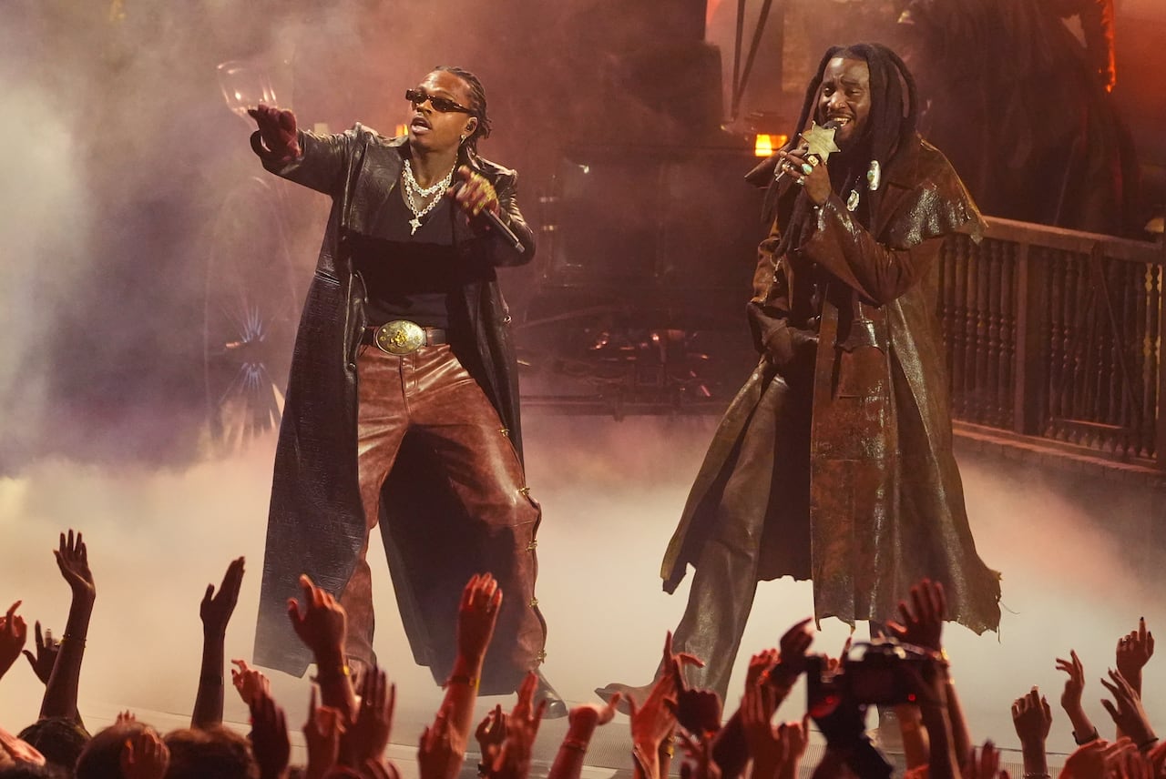Two men in long coats perform on a smoky stage.