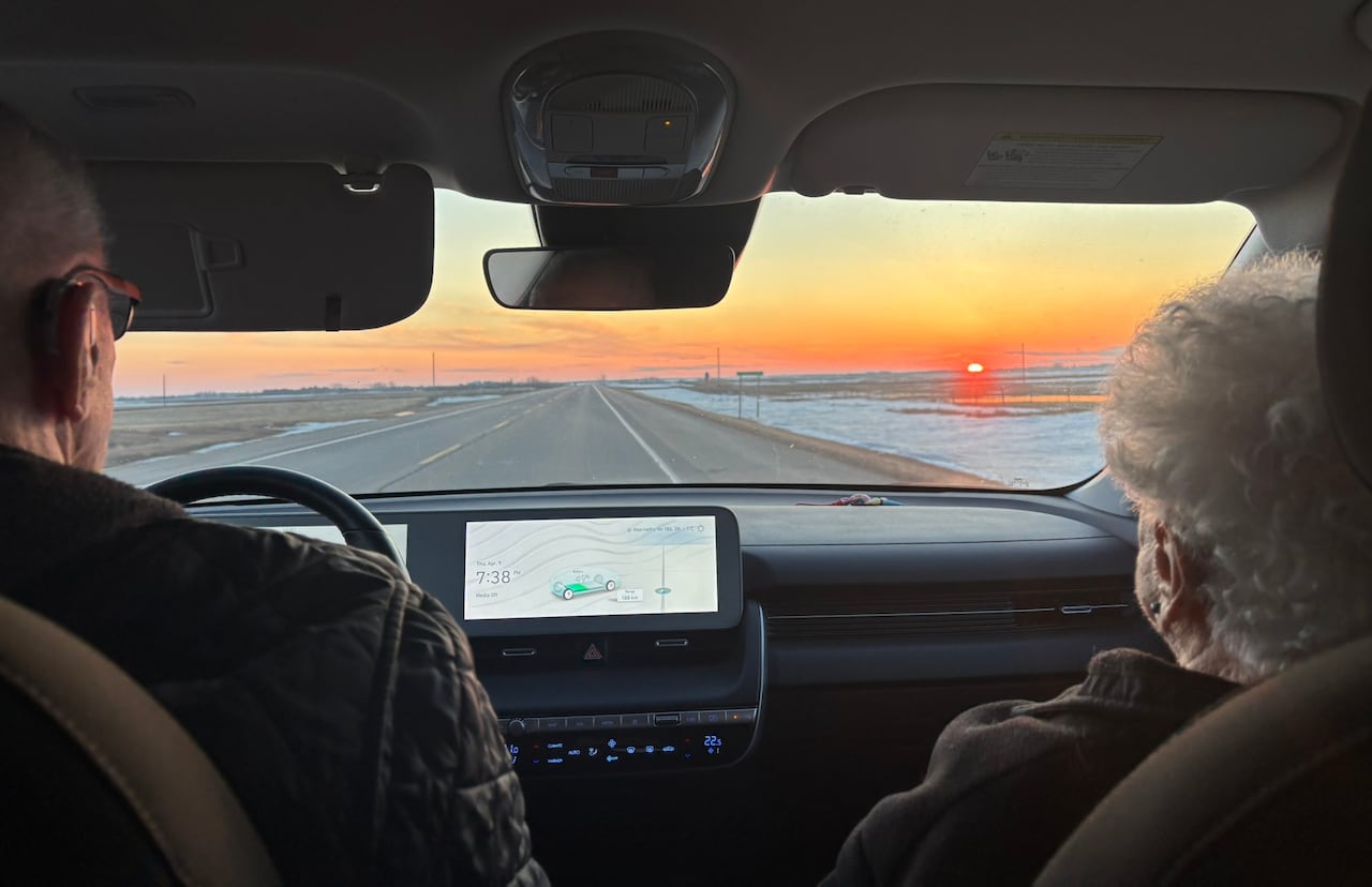 View from the back seat of a car with a man driving and a woman in the passenger seat. A highway, snowy fields and a sunset can be seen through the windshield.