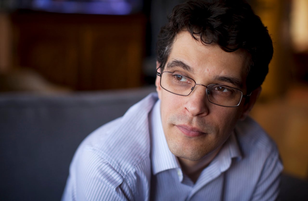 A man in spectacles and a light blue, striped shirt is seen in closeup, looking off to the left of the frame.