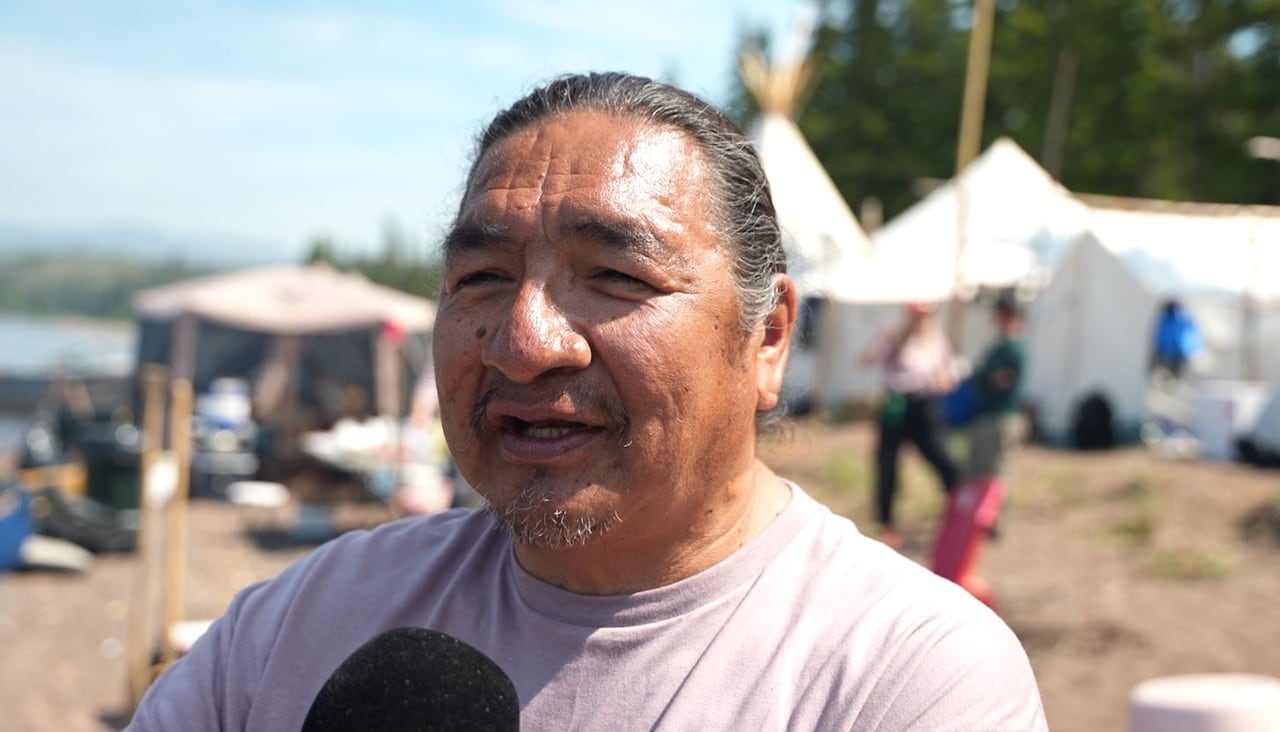 An Innu man with his hair tied back is pictured in front of a busy beach, with a variety of Innu tent structures in the background.