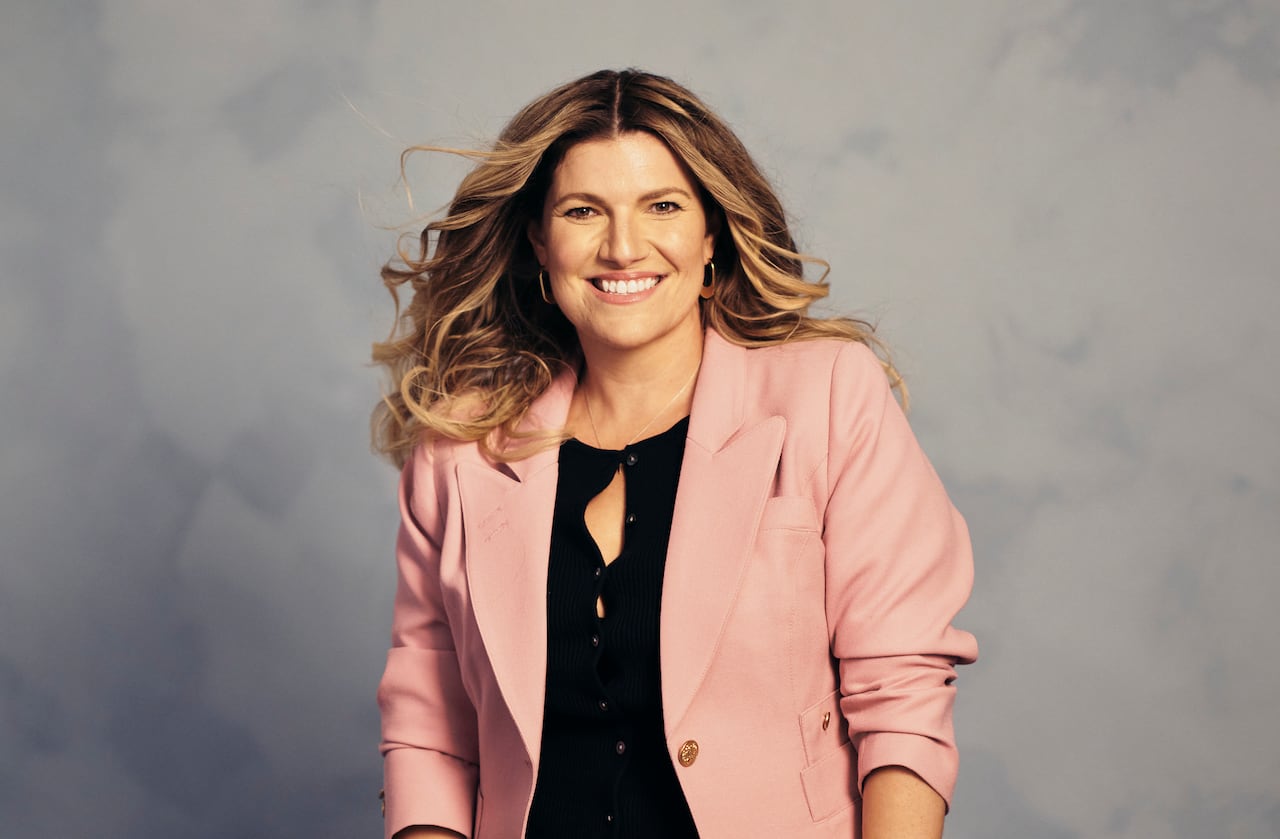 A woman in a pink blazer and blue jeans stands in front of plain backdrop.