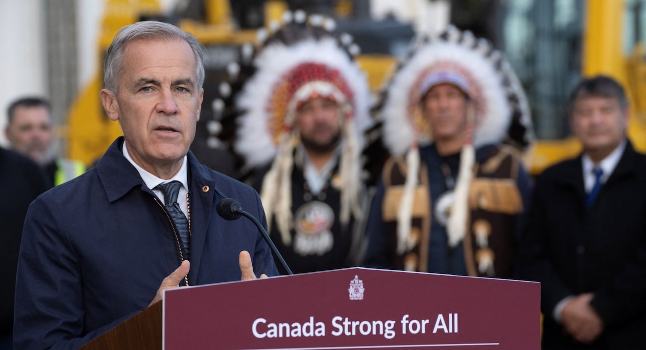 The Prime Minister speaks at a podium with two Indigenous leaders shown among a crowd in the background.