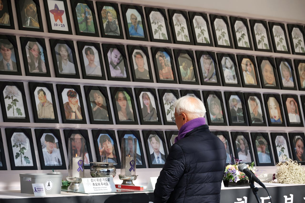 An elderly man prays in front of a wall showing dozens of photo portraits of mostly young people.