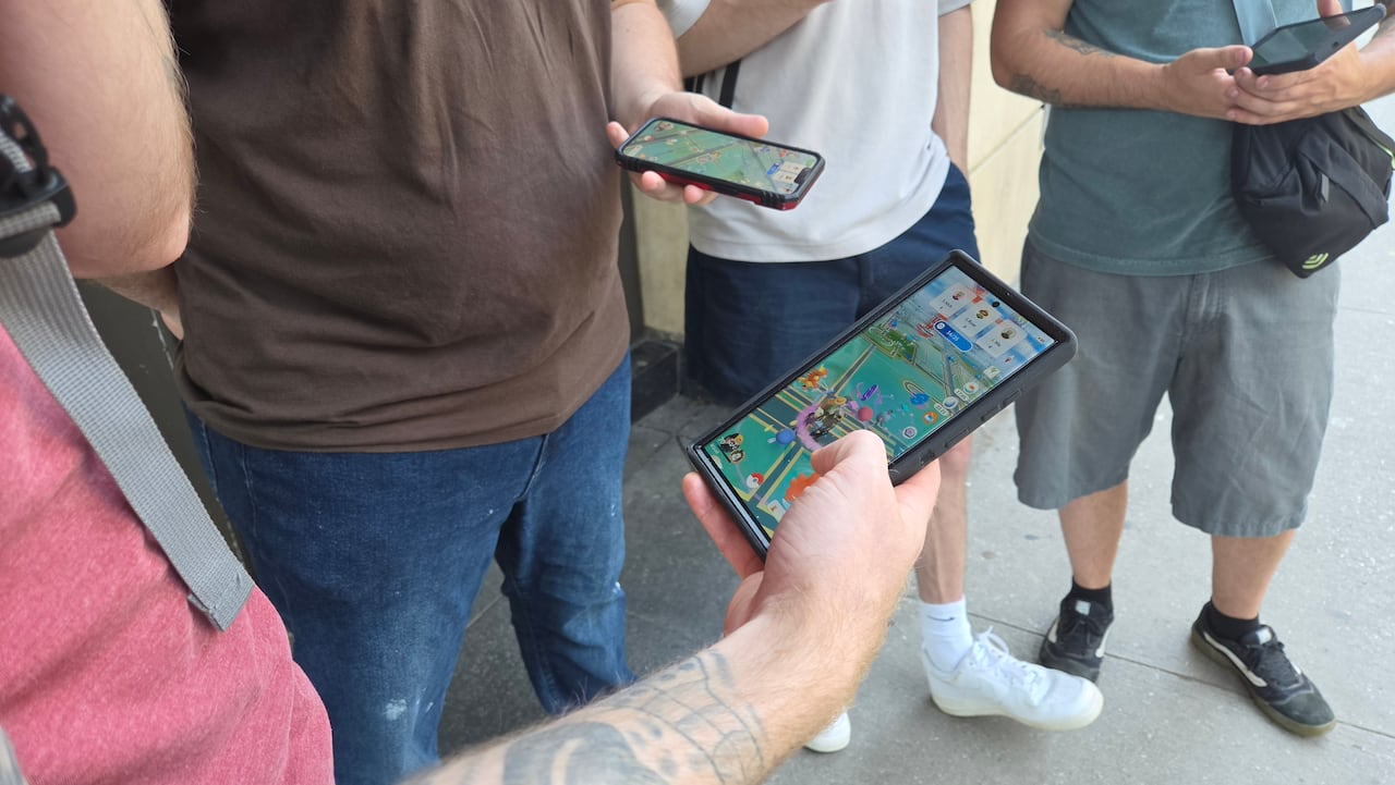 People's bodies and hands are seen holding phones that are playing a Pokemon game.