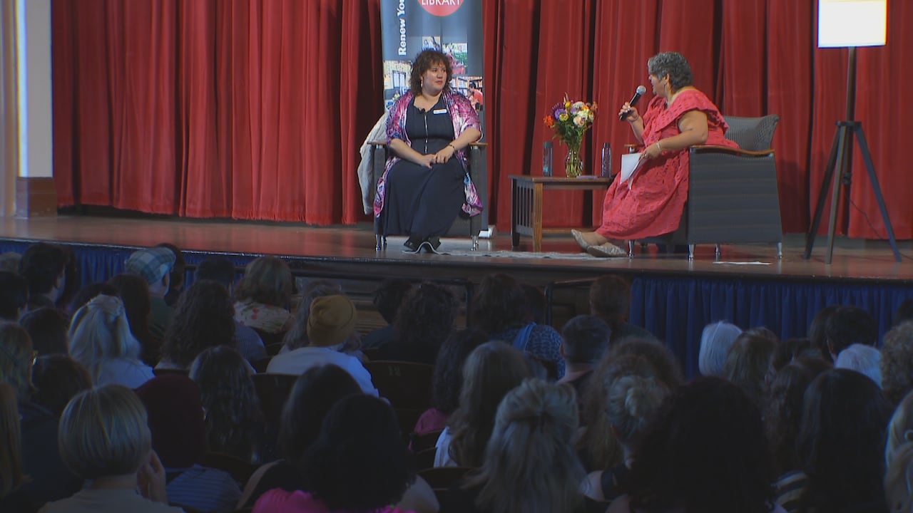 TWO PEOPLE IN CHAIRS, IN FRONT OF AN AUDIENCE