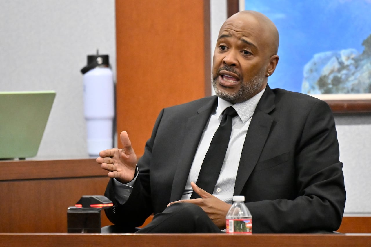 A man in a black suit, white shirt and black tie talks at a stand in a courtroom.