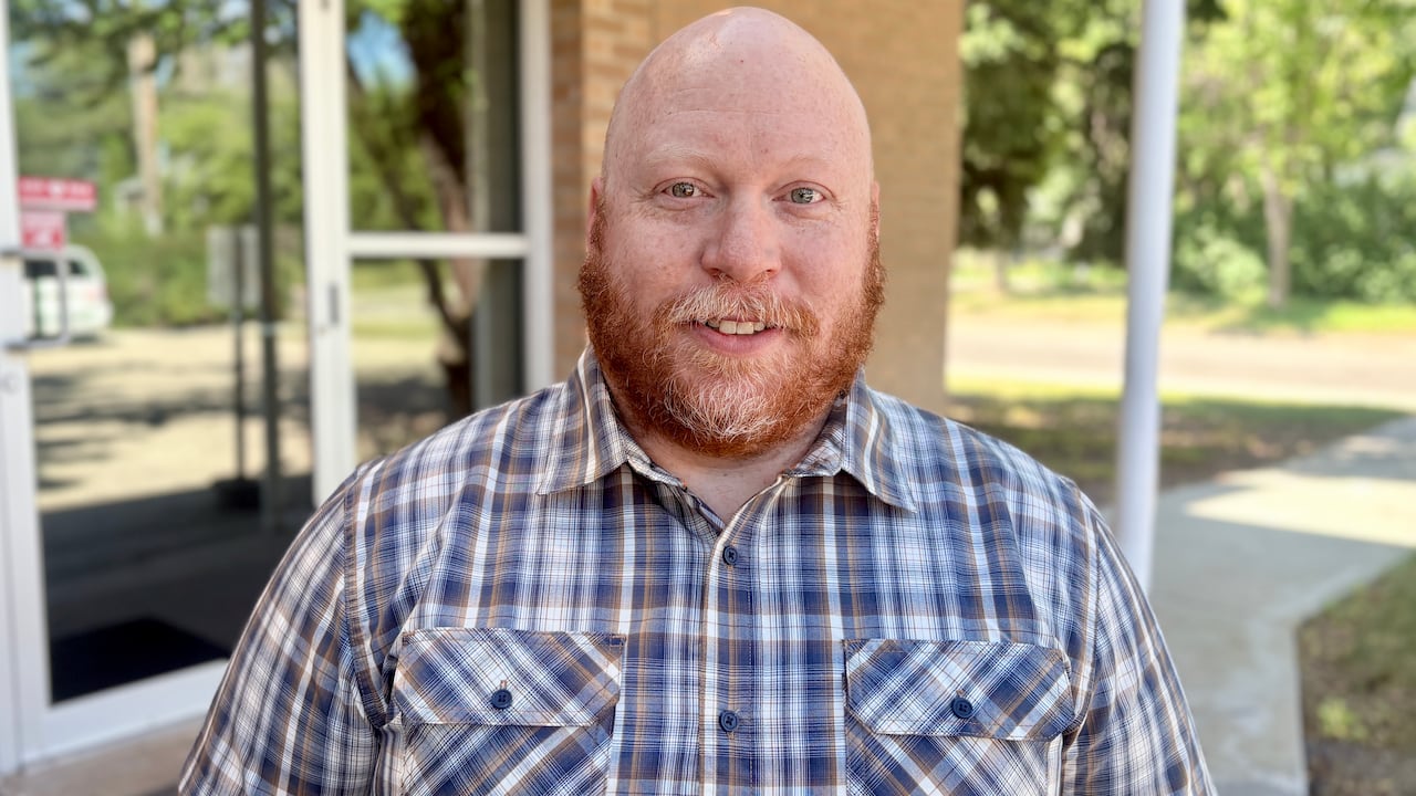 A man with a red beard stands outside a building on a sunny summer day.