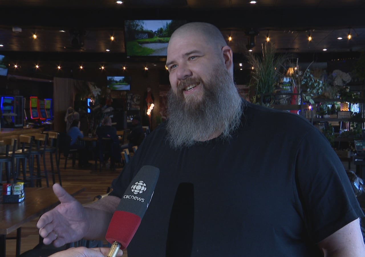 A bald man with a grey beard and a black shirt stands in a dimly lit bar.