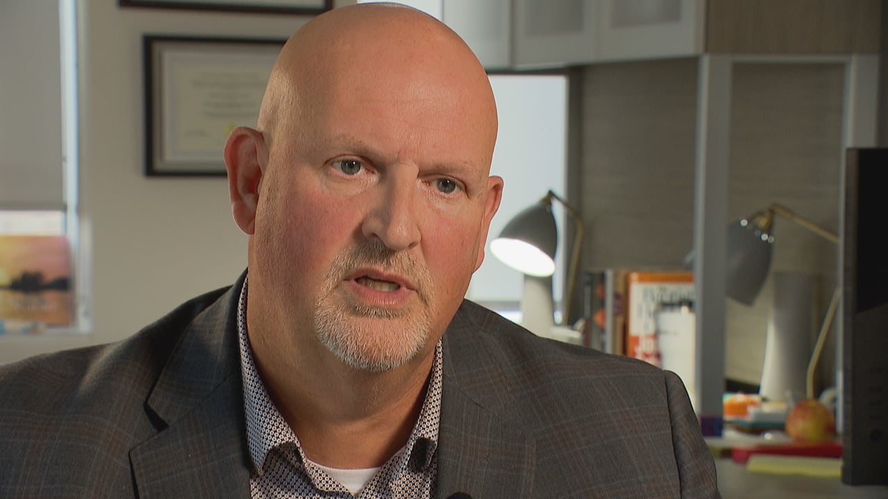 A seated man with a grey goatee and wearing a suit jacket and dress shirt speaks inside an office, a computer desk seen behind him.