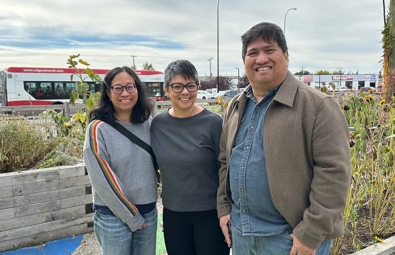 Three people stand together in a garden in front of a road.