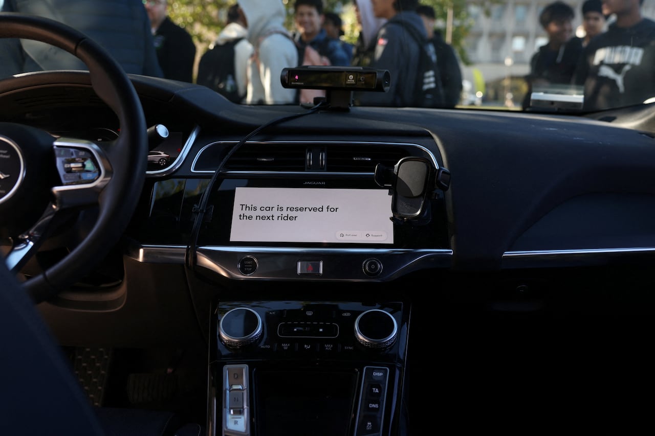 The all black interior of a Waymo without a driver at the wheel is depicted while the vehicle waits for its next passenger. On the screen the display reads "this car reserved for the next the rider."