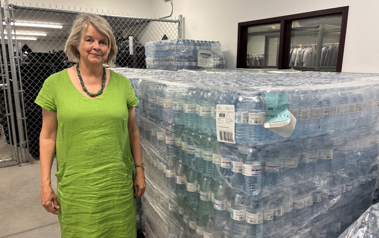A woman in a long green dress smiles and stands next to large pallets full of plastic water bottles.
