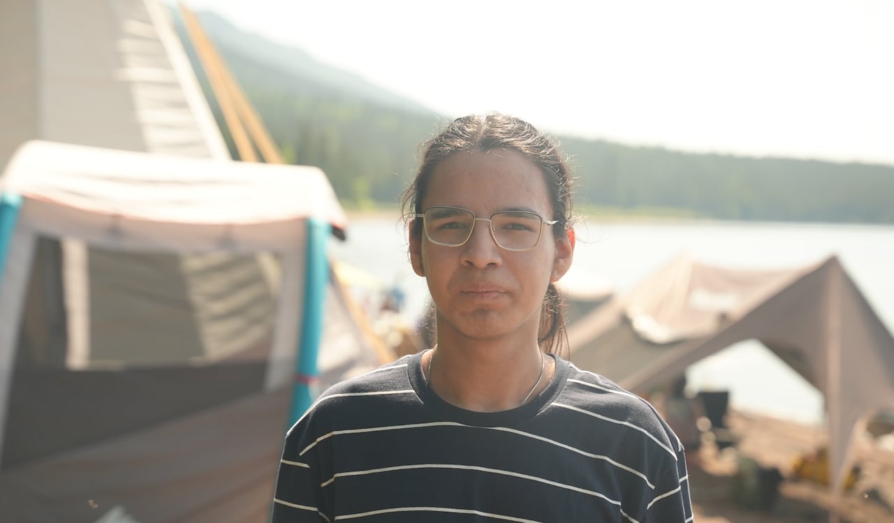 An Innu teen from Sheshatshiu is pictured in front of a traditional Innu tent. He has long, dark hair and wears glasses.