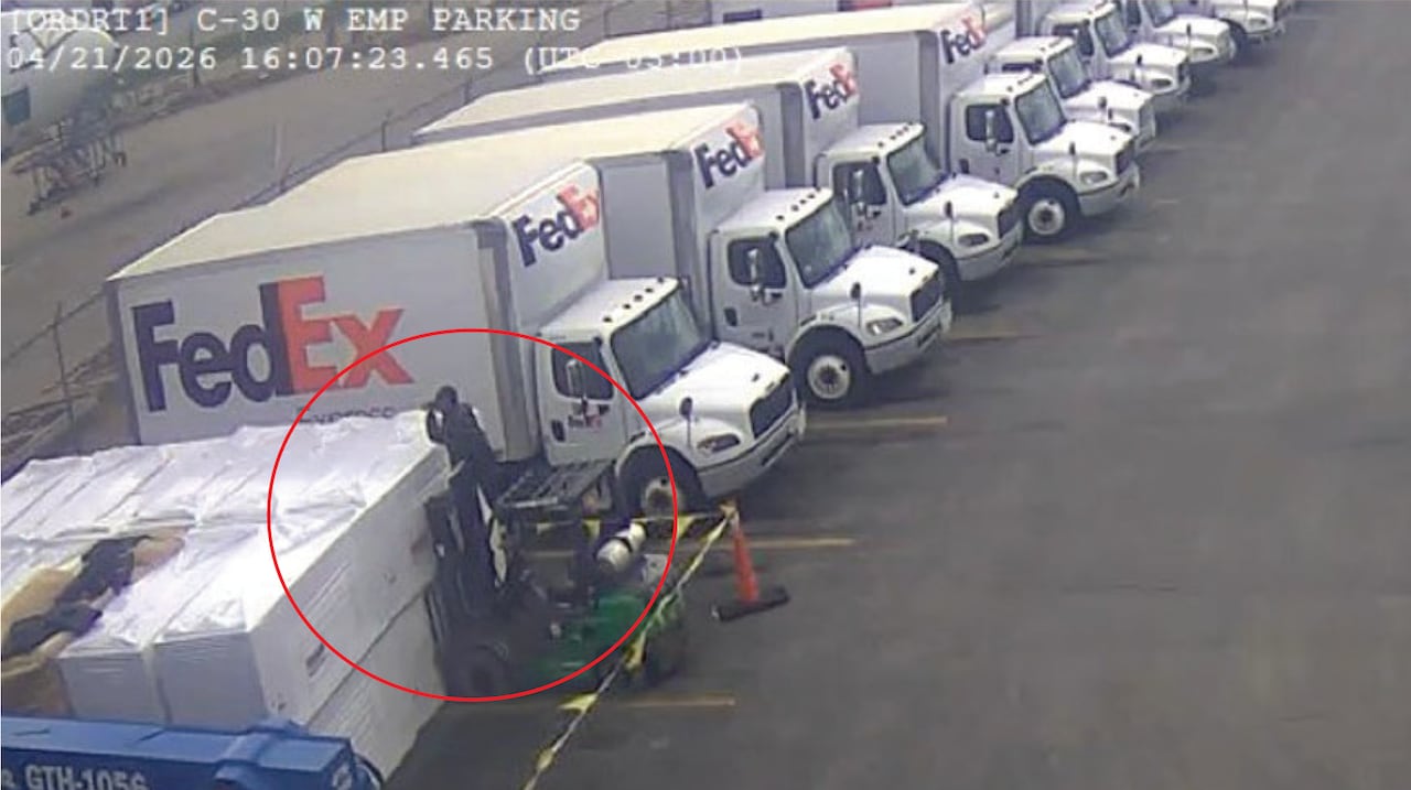 A man holds a camera while standing on a parked forklift, next to a row of FedEx trucks