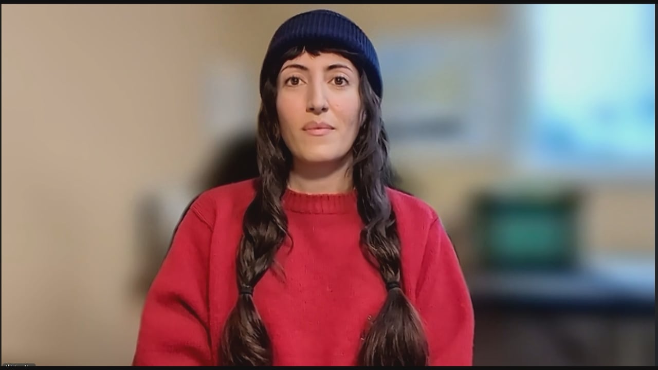 A woman in a beanie and a red sweater sits in front of a blurred background.
