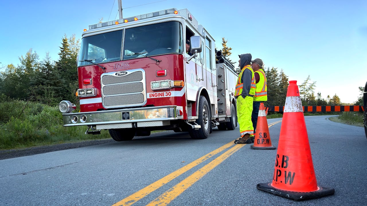 A fire truck is parked on the side of a road that's been blocked off by an evacuation order. Two individuals in safety suits speak to the driver outside their window.