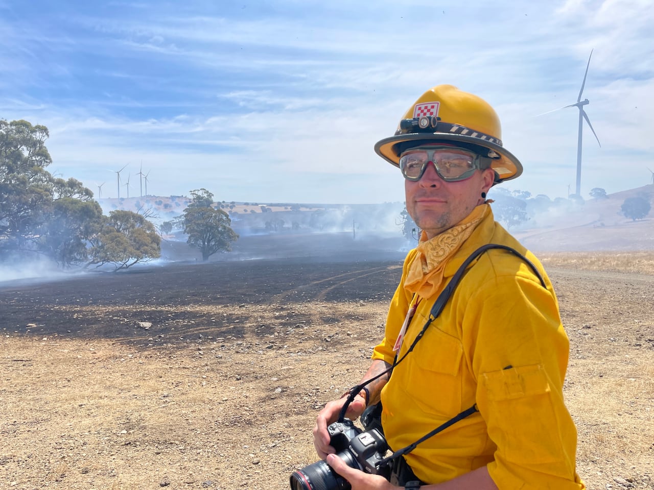Man in yellow firefighting suit, goggles and camera stands in front of smoke and wind turbines